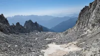 a debris covered glacier is framed by rocky walls on either side. In the distance are a series of ridges and valleys silhoutted. the sky is clear and blue.